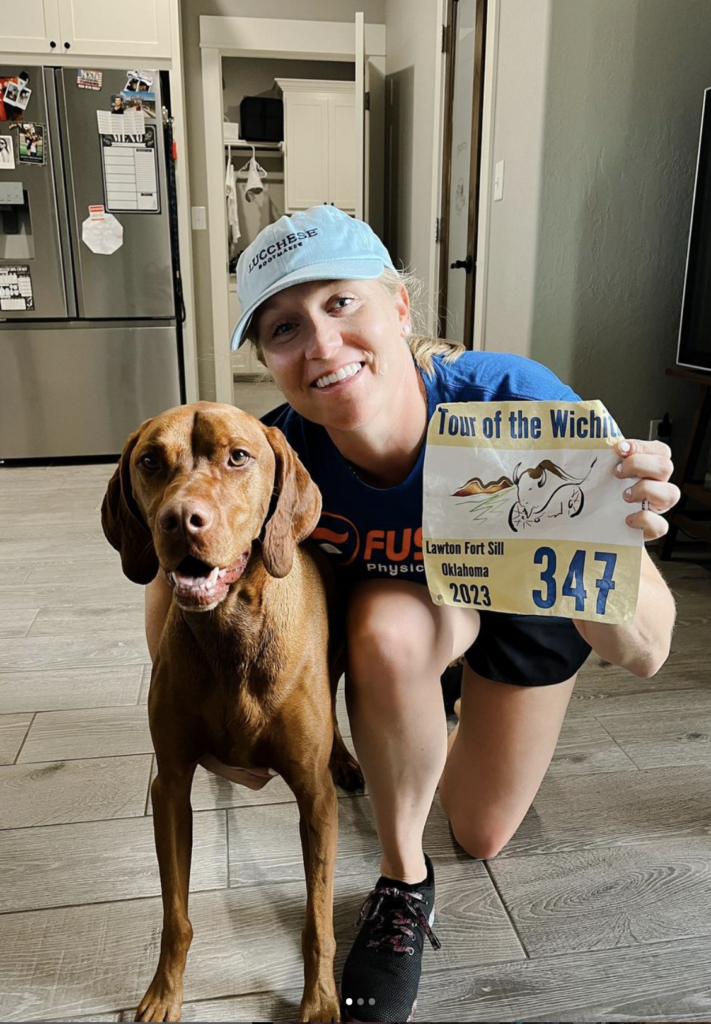 girl wearing blue hat showing race bib and smiling next to a brown dog